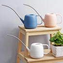 Modern April Showers Garden Watering Can placed on a wooden shelf surrounded by houseplants and flowerpots.
