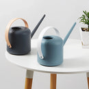 White and grey modern garden watering can surrounded by houseplants in a domestic setting.