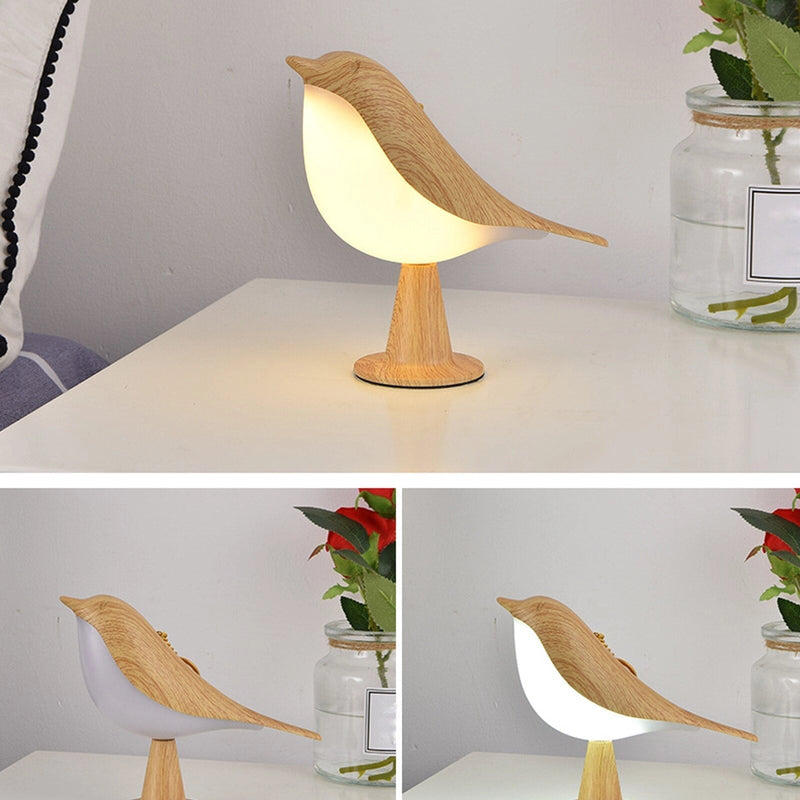 Dimmable LED cordless table nightstand light designed as a bird, placed on a wooden table next to a white flowerpot with houseplants. The lamp emits a soft orange and yellow glow, enhancing the cozy interior design.