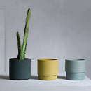 Bright porcelain stackable flowerpot planters featuring a variety of houseplants, including flowering plants in cylindrical and rectangular shapes, set on a wooden surface.