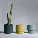 Stackable porcelain flowerpot planters with a variety of houseplants and flowering plants in cylindrical and rectangular shapes, set on a wooden surface.