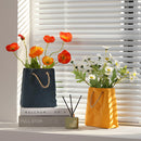 Ceramic vase designed to look like a shopping basket, featuring orange and yellow flowers and petals, placed near a window, enhancing the interior design.