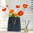 Ceramic vase with a shopping basket design featuring an arrangement of orange flowers and plants, set against an interior design backdrop with a window.