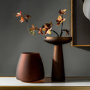 Coffee-colored frosted glass vase with a twig and a flower on a wooden table, illuminated by soft lighting.