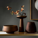 Coffee-colored frosted glass vase on a wooden table, surrounded by houseplants, twigs, and decorative items, enhancing the interior design.