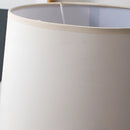 Contemporary beige lampshade table lamp featuring a metal base and wooden accents, placed next to drinkware and dishware on a linen-covered table in a living room or bedroom setting.