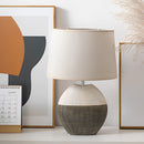 Contemporary lampshade table lamp made of plywood, featuring a rectangular design with artistic shading, displayed on a wooden shelf in a living room or bedroom setting.