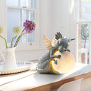 Dragon-shaped LED table lamp on a desk with purple and yellow accents, surrounded by plants and flowers near a window in an interior setting.