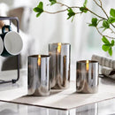 Three-piece LED pillar candles in a flameless glass candle set with remote control, displayed on a wooden table surrounded by plants, drinkware, and serveware, enhancing the interior design.