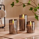 Three-piece flameless LED pillar candle set displayed on a wooden surface, surrounded by plants and elegant drinkware, with ambient interior lighting.