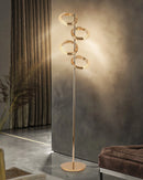 Gold diamond-patterned floor lamp with adjustable angles, featuring a wood base and twig-like sculpture near a glass table, complemented by a plant.
