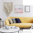 Handwoven macrame boho wall hanging tapestry with geometric art decor, displayed over a couch in a room with white furniture, a table, and a plant.