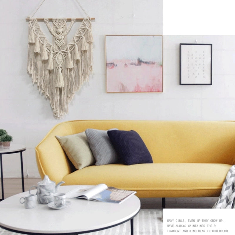 Handwoven macrame boho wall hanging tapestry with geometric art decor, displayed over a couch in a room with white furniture, a table, and a plant.