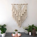 Handwoven Macrame Boho wall hanging tapestry featuring geometric art decor, displayed on a white wall with houseplants and wooden elements in an interior setting.