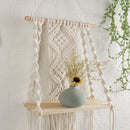 Beige handwoven macrame boho wall hanging tapestry with geometric patterns and embellishments, displayed on a wooden shelf.