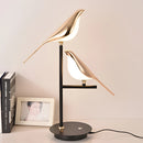 Postmodern LED desk lamp featuring a galvanized golden bird design, with elements of wood and metal, complemented by an artistic lampshade on a shelving unit.
