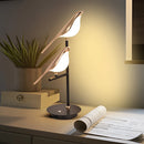 LED desk lamp featuring a postmodern galvanized golden bird design, placed on a wooden table surrounded by plants and office supplies, enhancing the interior design.