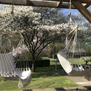 Macramé hammock swing hanging from a tree in a garden with lush green grass and a backdrop of the sky and various shades of green foliage.