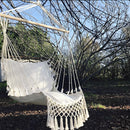 Macramé hammock swing hanging from a tree in a garden, surrounded by lush green plants and grass under a clear sky.