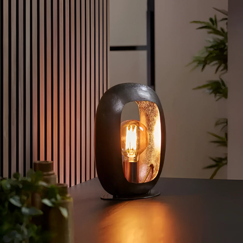 Small vintage-style metal table lamp with amber-colored lighting, featuring wooden elements and artistic design in shades of orange, placed next to a plant in an interior setting.