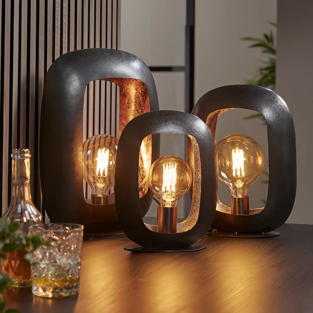 Vintage-style metal table lamp with intricate designs, displayed in a warmly lit interior setting, accompanied by glass and wooden decor elements.