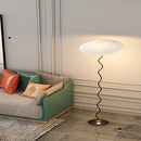 Curved pleated floor lamp with a metal textile shade, placed next to a couch on wooden flooring, enhancing the room's comfort and lighting.