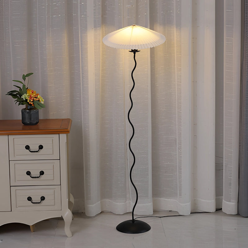Curved metal floor lamp with pleated textile shade, placed near wooden cabinetry and a plant in a stylishly designed interior.