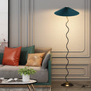 Curved pleated floor lamp with a metal and textile design, featuring a green and black color scheme, positioned on a wooden floor in a well-lit, comfortable interior setting.