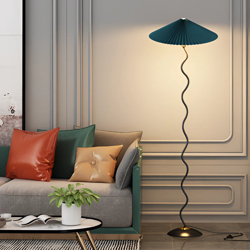 Curved pleated floor lamp with a metal and textile design, featuring a green and black color scheme, positioned on a wooden floor in a well-lit, comfortable interior setting.
