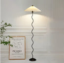 A curved pleated floor lamp with a metal textile shade, set on a wooden base.