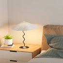 Curved pleated floor lamp with a metal and textile shade, set on a wooden floor next to a houseplant, enhancing a cozy and artistic home ambiance.