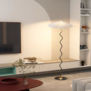 Curved pleated floor lamp made of metal and textile, situated in a living room with wooden flooring and furniture.