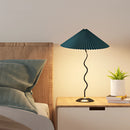 Curved pleated floor lamp made of metal and textile, placed beside a plant, in a cozy interior setting with wooden elements and soft lighting.