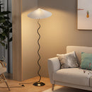 White metal textile curved pleated floor lamp with a shade, set in a cozy interior featuring wooden flooring and soft lighting.