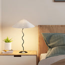 A curved pleated floor lamp made of metal and textile, featuring a shade, situated in a cozy interior with wooden elements, including a table and plant near a wall.