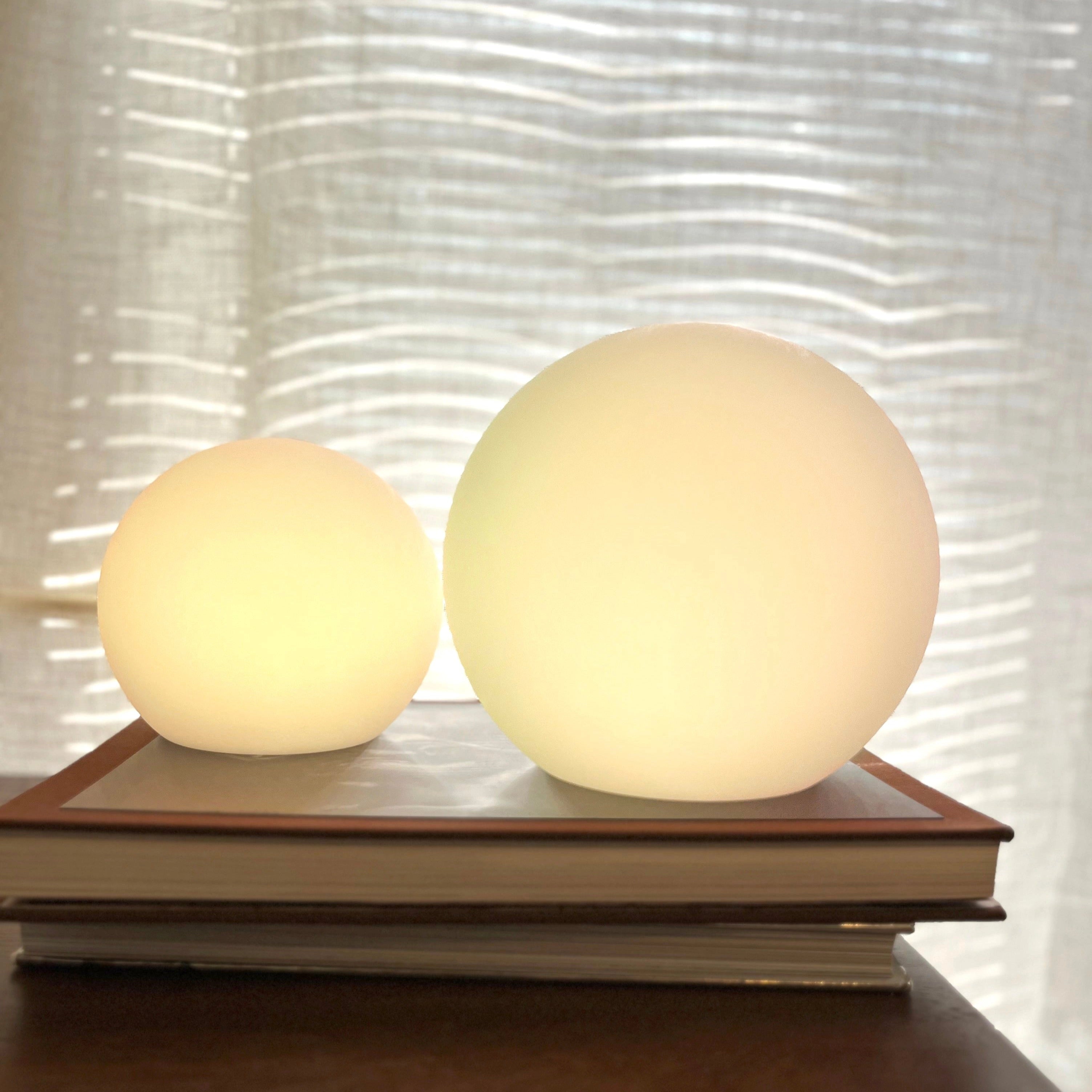 Cordless LED table lamp with color control settings, displayed on a wooden table, featuring a beige lampshade and artistic decor elements in the background.