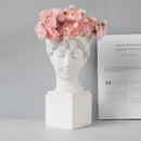 Neoclassical Greek head sculpture used as a vase, adorned with plants and flowers, featuring artful details and a headpiece-like design.