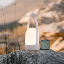 Rechargeable table lamp with touch dimming feature, illuminated and placed on a wooden surface against a wall, with a soft glow highlighting a winter landscape scene with trees and rocks in the background.