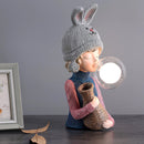 Elegant resin table lamp featuring a white rabbit girl statue with a dual glass lampshade, surrounded by decorative items including a wood fawn figure, a doll, and assorted fashion accessories.