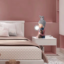 Resin Table Lamp Elegance Rabbit Girl with Dual Glass Lampshade featuring a detailed rabbit girl figurine, set in a cozy bedroom with grey pillows, a wooden bed frame, and soft textiles near a window.
