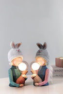 Decorative resin table lamp featuring an elegant rabbit girl design with a dual glass lampshade, accented with grey and fawn tones, and adorned with woolen and wooden artistic elements.