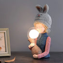 Resin Table Lamp Elegance Rabbit Girl with Dual Glass Lampshade featuring a human-like figure with knee-length attire and headgear, positioned near a wooden picture frame under soft lighting.