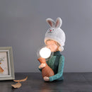 Resin table lamp featuring an elegant rabbit girl design with a dual glass lampshade, surrounded by items including a wood frame and various textured fabrics.