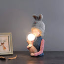 Resin table lamp designed as an elegant rabbit girl, featuring dual glass lampshades, placed on a wooden floor, surrounded by artistic and musical elements in a well-decorated room.