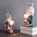 Resin table lamp featuring an elegant rabbit girl design with dual glass lampshades, surrounded by artful decor and a cozy room setting.