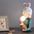 Resin table lamp featuring a design of a rabbit girl in costume with a dual glass lampshade, surrounded by elements of wood and art, giving a happy and elegant ambiance.