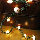 Battery-operated LED garland featuring amber rose flower string lights intertwined with green leaves and branches, displayed on a wooden wall, enhancing the interior design with soft lighting.