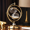 Elegant sand clock designed as a rotating globe hourglass timer, featuring a metallic and antique appearance with elements resembling automotive lighting and headlamp designs, displayed in a classic still life setting.