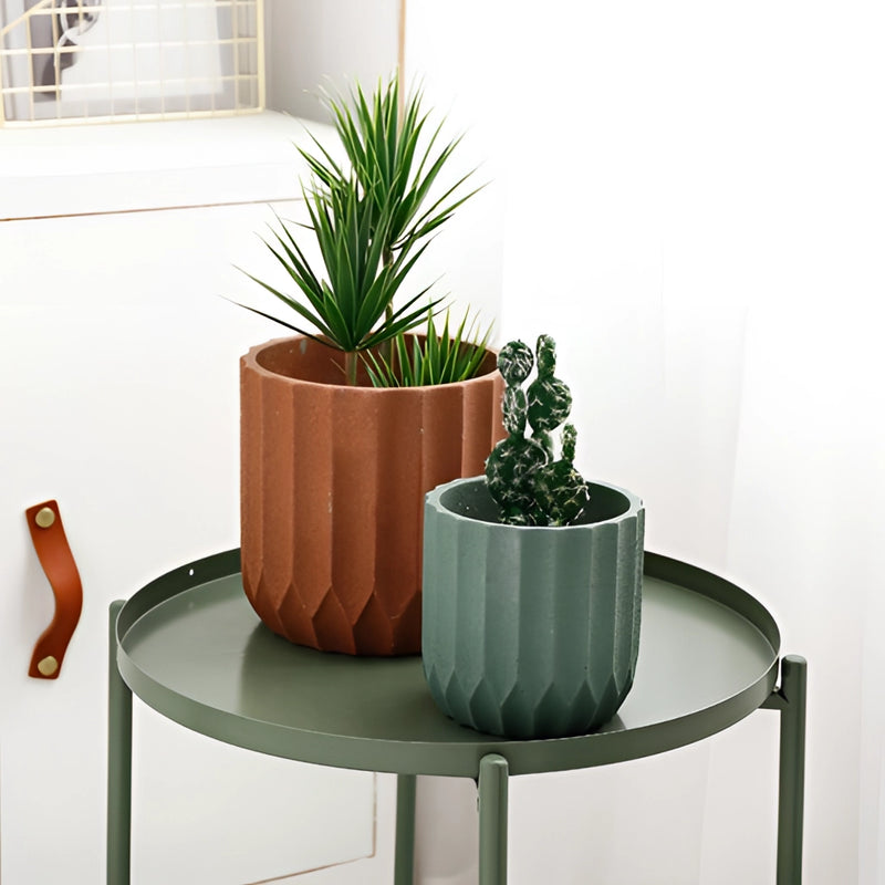 Textured ceramic polygon vase with a geometric design, placed on a wooden floor near a window, surrounded by houseplants in an interior setting.