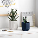 Textured ceramic polygon vase with a geometric design, displayed on a table alongside houseplants, in a well-lit interior setting with a grey couch.